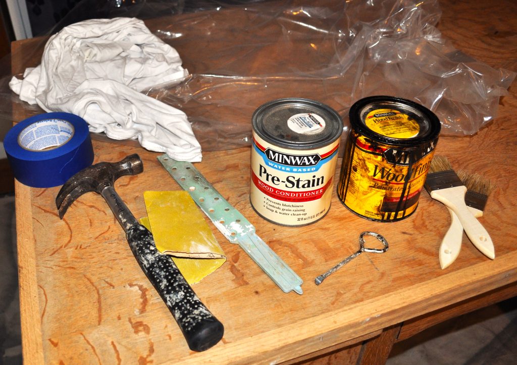 Supplies on a wooden unfinished desk. Rags, plastic, painters tape, paint stir stick, hammer, sandpaper, paint can opener, paint brushes, pre-stain, stain.
